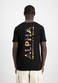 Svart bomull t-skjorte med en vertikal flerfarget grafikk som leser "ALPHA INDUSTRIES" på ryggen. Vanlig passform med korte ermer.