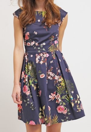 Ärmelloses marineblaues Kleid mit floralem Muster, tailliertem Schnitt und plissiertem Rock, getragen von einer Person mit langen braunen Haaren.