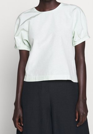 Personne à la peau foncée portant un haut blanc ample à manches courtes bouffantes et un pantalon noir, vue du cou aux hanches.