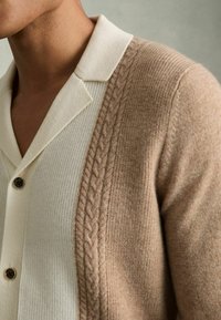 Cardigan en maille beige avec un motif de tricot en câble d'un côté, associé à une chemise blanche à côtes dotée d'un col et de détails de boutons.