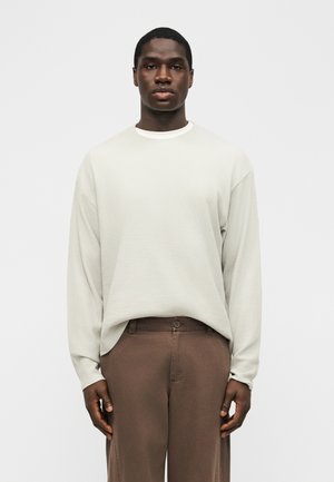 Νεαρός άνδρας που κοιτάζει προς τα εμπρός, φορώντας μια χαλαρή, ανοιχτό μπεζ μακρυμάνικη μπλούζα, μπουρντουρισμένη μέσα σε καφέ παντελόνια ψηλής μέσης, μπροστά σε απλό φόντο.