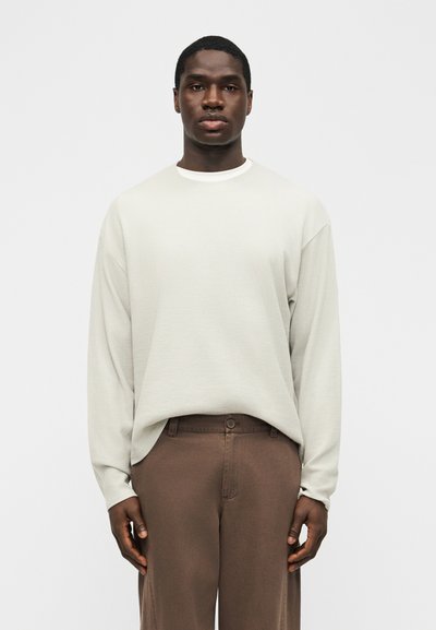 Jeune homme de face portant une chemise ample à manches longues beige clair rentrée dans un pantalon marron taille haute, sur un fond uni.