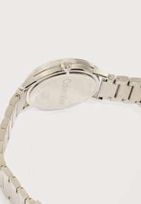 Ασημένιο ανδρικό ρολόι χειρός Calvin Klein από ανοξείδωτο ατσάλι που δείχνει το πίσω κάλυμμα με ανάγλυφο το λογότυπο της μάρκας και μεταλλικό μπρασελέ με κρίκους, σε λευκό φόντο.