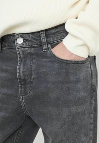 Jean en denim gris foncé avec un design classique à cinq poches et une fermeture à bouton métallique. Le tissu présente une légère texture abîmée.