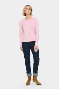 Cardigan rose clair avec une texture côtelée, col rond, fermeture à boutons sur le devant, porté sur un jean en denim foncé et des bottines brunes.