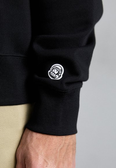 Sweatshirt noir avec un poignet côtelé et un patch rond blanc représentant le visage d'un astronaute sur la manche.