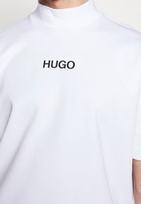 Personne portant une chemise blanche à col montant avec des manches courtes et le mot « HUGO » imprimé en noir, centré sur la poitrine.