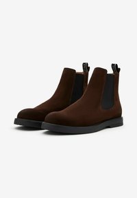 Bruine suède Chelsea boots met zwarte elastische zijkanten, een ronde neus en een zwarte rubberen zool. Voorzien van een trekband voor eenvoudig aan- en uittrekken.