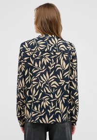 Chemise à manches longues en tissu bleu marine, ornée d'un motif de feuilles beige clair. Col classique, dos à boutons avec un ourlet droit.