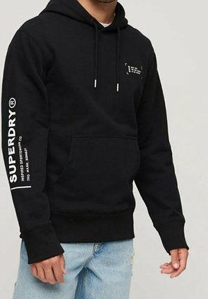 Mężczyzna noszący czarną bluzę z kapturem Superdry z białym napisem na lewym rękawie i klatce piersiowej, w połączeniu z jasnoniebieskimi jeansami.