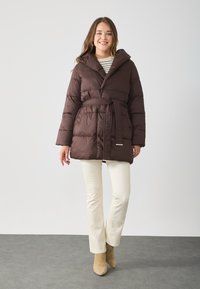 Brun pufferjakke med belte i midjen, hette og quiltet design. Kombinert med kremfargede bukser og beige ankelstøvler på en grå gulv.