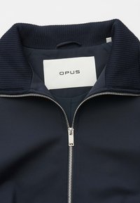 Veste bleu marine avec col côtelé et fermeture éclair argentée, affichant l'étiquette de la marque "OPUS" à l'intérieur du col.