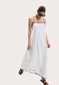 Bílé maxi šaty se střihem do rovna, černým lemováním podél výstřihu a lemu a tenkými ramínky. Modelka doplněná tmavými slunečními brýlemi a sandály.