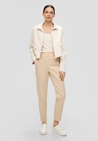 Veste courte beige clair avec deux poches avant, associée à un pantalon beige clair et des baskets blanches. Tissu lisse et design minimaliste.