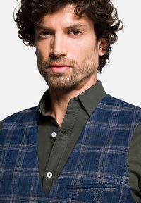 Granatowa kamizelka w kratę z teksturowanej tkaniny na ciemnozielonej koszuli z guzikami. Kamizelka ma jedną kieszeń na piersi i białe guziki.