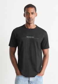 Juodas medvilnės marškinėliai trumpomis rankovėmis, su „Calvin Klein Jeans“ logotipu baltu, apvalia iškirpte ir laisvu kirpimu.