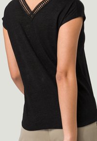 Kvinna som bär en svart kortärmad topp med en mesh V-ringning och beige byxor, visad från midjan till mitten av låret.