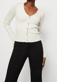 Kremowy kardigan z prążkowanej dzianiny z dekoltem w kształcie litery V, z zapięciem na guziki i długimi rękawami. Połączony z dopasowanymi czarnymi spodniami.