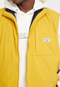 Gelbe Nylonweste mit Reißverschluss vorne, einer Brusttasche und lockerem Schnitt, getragen über einem weißen Hoodie mit bedrucktem Text.