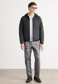 Vīrietis, kurš ģērbies melnā quilts jaku, baltā kreklā, pelēkās biksēs, melnās sporta kurpēs un saulesbrillēs, stāv pret vienkāršu baltu fonu.