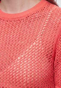Gros plan d'une personne portant un pull à maille ajourée corail par-dessus un top uni assorti, montrant un tissu texturé et transparent.
