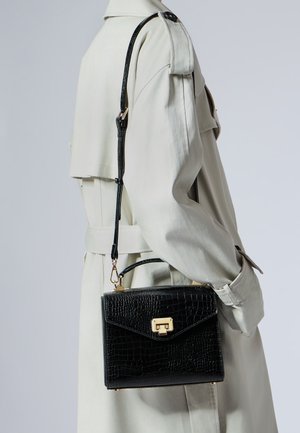 Sac à main en cuir noir texturé avec fermoir doré et bandoulière porté sur un trench crème sur le côté droit du torse.