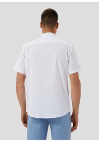 Uomo che indossa una camicia bianca a maniche corte e pantaloni azzurri chiari, in piedi con la schiena rivolta verso l'osservatore su uno sfondo grigio chiaro.