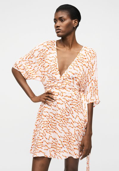 Γυναίκα που φοράει ένα λευκό φόρεμα τύπου wrap με πορτοκαλί λουλουδάτο σχέδιο, μανίκια μέχρι τον αγκώνα και βαθύ V-λαιμόκοψη, στέκεται με το ένα χέρι στη μέση της.