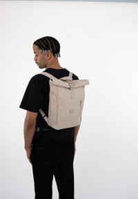Beige roll-top backpack with adjustable straps, external zip pocket, made of durable fabric, featuring a sleek, minimalist design.