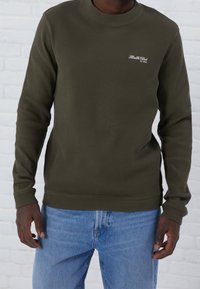 Suéter de manga larga verde oliva con textura acanalada, cuello redondo y logotipo bordado de "Health Club" en el pecho; combinado con jeans azules.