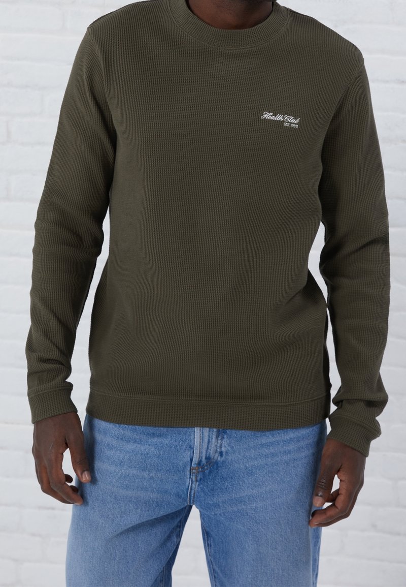 Suéter de manga larga verde oliva con textura acanalada, cuello redondo y logotipo bordado de "Health Club" en el pecho; combinado con jeans azules.