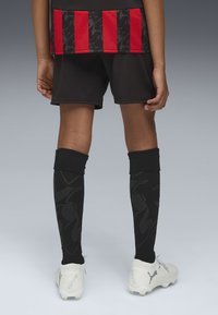 Schwarze Fußballshorts werden mit einem rot-schwarz gestreiften Trikot kombiniert, das ein dezentes Muster aufweist. Schwarze Socken mit einem Design und weiße Fußballschuhe runden den Look ab.