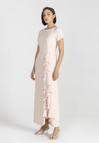 Długa, bladoróżowa sukienka z krótkimi rękawami i falbanką z przodu. Wykonana z gładkiego materiału, z dopasowaną górą i rozkloszowanym dołem.