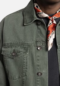 Olijfgroene spijkerjas met twee borstzakken met knoopsluitingen, klassieke kraag en subtiele textuur, gecombineerd met een zijden sjaal met patroon.