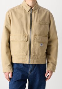 Beżowa bawełniana kurtka z zamkiem błyskawicznym, dwoma przednimi kieszeniami i małą kieszonką na piersi. Posiada stójkę i prostokątną metkę z logo.