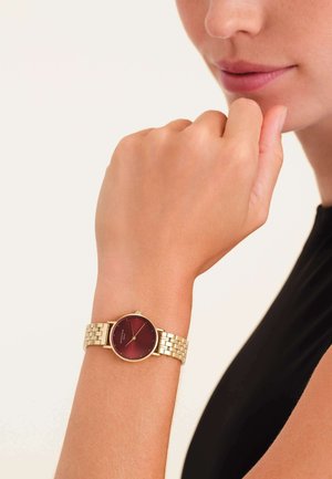 Montre-bracelet en métal doré avec un cadran rond couleur bordeaux et des maillons dorés assortis. La montre est portée sur une main dans une pose naturelle et subtile.