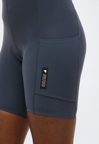 Κοντινό πλάνο σφιχτών αθλητικών σορτς σκούρου γκρι με πλαϊνή τσέπη και μαύρη ετικέτα με λευκό κείμενο στο μηρό ενός ατόμου.