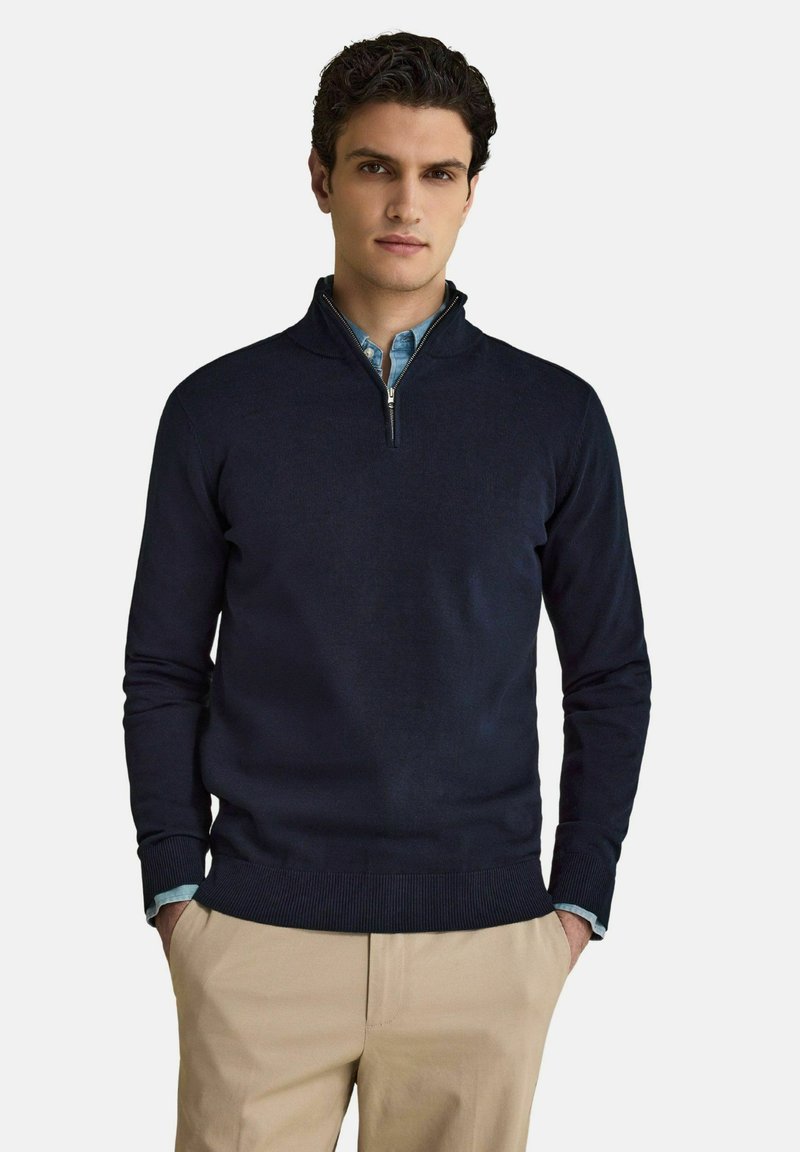 Homme aux cheveux bouclés et foncés portant un pull bleu marine à fermeture éclair quart, par-dessus une chemise bleu clair et un pantalon beige, debout les mains dans les poches.