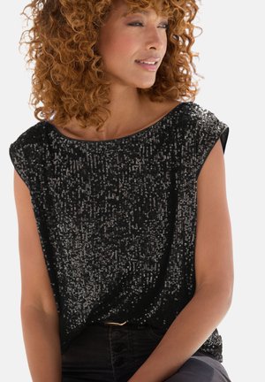 Frau mit lockigem Haar, die ein ärmelloses schwarzes Top trägt, bedeckt mit kleinen glänzenden Pailletten, leicht nach rechts gewandt mit einem sanften Lächeln.