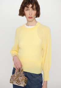 Hellgelber Strickpullover mit rundem Halsausschnitt und langen, leicht puffigen Ärmeln. Kombiniert mit einer kleinen, verzierten Samt-Handtasche.