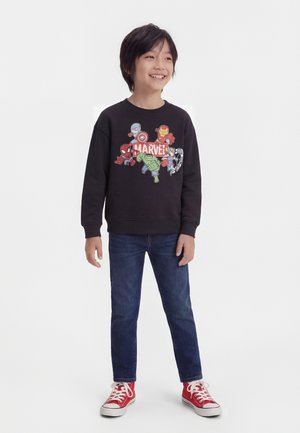 Garçon souriant debout, portant un sweat-shirt Marvel noir avec Spider-Man, Captain America, Iron Man, Hulk et Thor, un jean bleu et des baskets rouges.