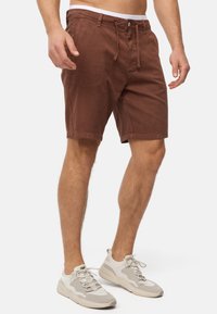Shorts en coton marron avec une coupe décontractée, cordon de serrage et fermeture à bouton, poches avant et bords ourlés. Associés à des baskets gris clair.