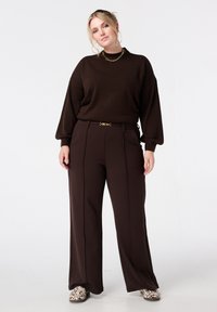 Bruine oversized trui met lange mouwen, gecombineerd met een high-waist, bruine wijde pijpen broek met een gouden gesp riem en patroon schoenen.