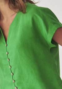 Blusa in lino verde con scollo a V, maniche corte arrotolate e una fila di sette bottoni in legno lungo la patta frontale. Tessuto liscio.