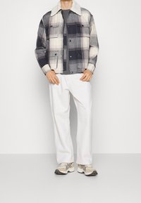 Kraciasta kurtka z kołnierzem sherpa w kolorach szarym, granatowym i kremowym, zestawiona z szarą koszulką i białymi, luźnymi spodniami.