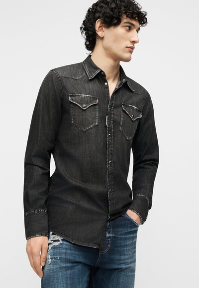 Νεαρός με σγουρά μαλλιά που φοράει ξεβαμμένο μαύρο τζιν πουκάμισο με κουμπωτές τσέπες στο στήθος και φθαρμένο μπλε τζιν, κοιτάζοντας προς τα πλάγια.