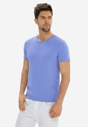 Uomo in piedi che guarda a destra, indossa una t-shirt aderente celeste a maniche corte e pantaloni bianchi su uno sfondo uniforme.