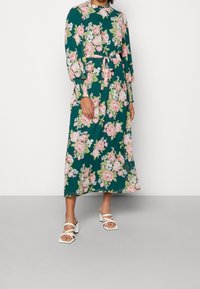 Vestido maxi floral con mangas largas, tela verde, patrones de flores rosas y blancas, cintura ajustada con un cinturón de lazo, combinado con sandalias blancas de tacón bloque.