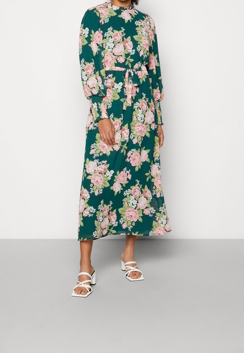 Vestido maxi floral con mangas largas, tela verde, patrones de flores rosas y blancas, cintura ajustada con un cinturón de lazo, combinado con sandalias blancas de tacón bloque.