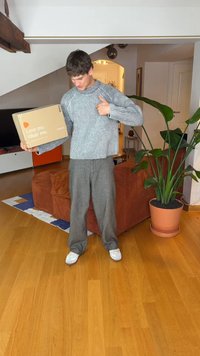 Striped knit sweater in grey tones, loose-fitting grey trousers, and white sneakers; holding a beige box labeled “Love me. Wear me.”